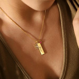 May include: A gold necklace featuring a rectangular pendant engraved with the name "Claudia" and the numbers "2025". The necklace also includes a small pink gemstone and a charm. The chain is delicate.