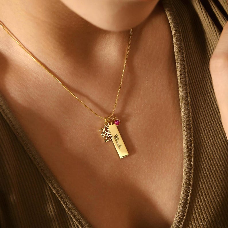 May include: A gold necklace featuring a rectangular pendant engraved with the name "Claudia" and the numbers "2025". The necklace also includes a small pink gemstone and a charm. The chain is delicate.