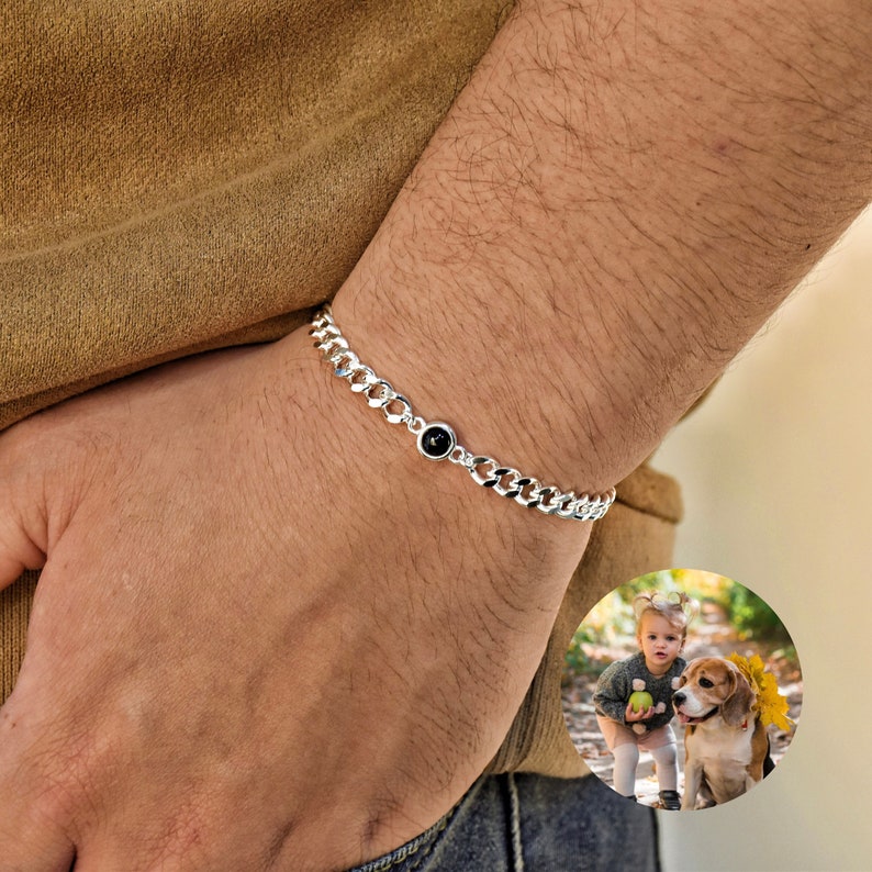 May include: A silver curb chain bracelet with a black gemstone center. The bracelet is worn on a wrist, showcasing its polished links and the dark, circular stone. The image includes a small inset photo of a child and a dog.