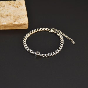May include: Silver chain bracelet with a small, round, dark-colored charm. The bracelet features a curb chain design and an adjustable chain closure. The background is black with a light-colored stone.