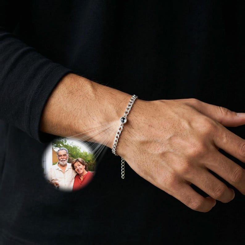 May include: A silver chain bracelet with a small, dark-colored stone. The bracelet is on a person's wrist. A small, circular image of a couple is projected onto the person's arm. The bracelet has a simple, elegant design.