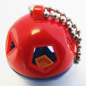 Puede incluir: Una bola de rompecabezas roja y azul con cortes geométricos. La bola tiene una cadena plateada. Dentro de los cortes hay formas azules y naranjas. El juguete está diseñado para desafiar al usuario a resolver el rompecabezas.