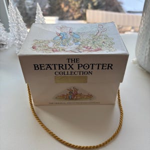 Peut inclure: Une boîte blanche avec une poignée en corde dorée. La boîte présente une image de Peter Rabbit sur le dessus et le texte "The Beatrix Potter Collection" sur le devant.