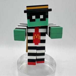 Puede incluir: Una figura de estilo Minecraft de un personaje con una cara verde, un cuerpo a rayas blancas y negras y un sombrero negro con una raya amarilla. El personaje tiene una corbata roja con un logotipo "M" dorado y manos verdes.