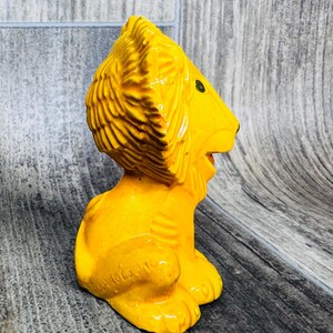 May include: A yellow ceramic lion figurine with a detailed mane and green eyes. The lion is sitting with its head turned to the side and its mouth slightly open.