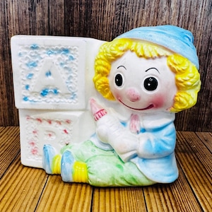 May include: A vintage ceramic baby figurine with a blue hat and dress, holding a bottle. The figurine is next to white blocks with the letters "A" and "B" on them. The figurine has yellow hair and is sitting on a green base.