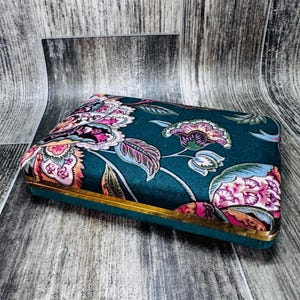 May include: A teal blue velvet box with a gold trim and a floral pattern. The box has a hinged lid and is decorated with pink, white, and green flowers.