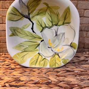 May include: Square ceramic bowl with a hand-painted floral design. The bowl features a white magnolia flower with yellow accents and green leaves. The rim is slightly curved, and the background is a woven texture.