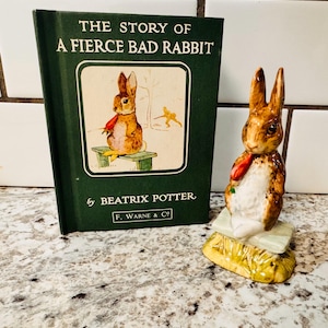May include: A hardcover book titled "THE STORY OF A FIERCE BAD RABBIT" by Beatrix Potter, next to a ceramic figurine of a rabbit holding a carrot. The book has a green cover with a rabbit illustration. The figurine is brown and white.