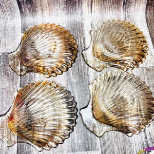 May include: Four decorative glass seashell-shaped dishes. The dishes are clear with a golden-brown hue, and feature a detailed ribbed texture. The dishes are arranged on a gray and white wooden surface.