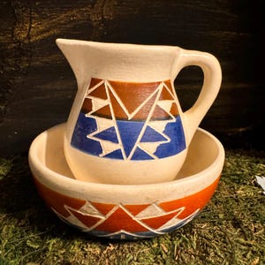 May include: A white ceramic pitcher and bowl set with a geometric pattern in shades of blue, red, and brown. The pitcher has a handle and the bowl is round.