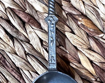May include: A silver-toned spoon with a detailed handle. The handle features a twisted design and a small sculpture of two figures at the top. The spoon rests on a woven, textured surface.