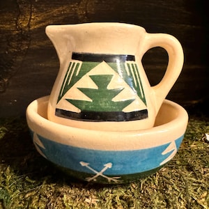 May include: A cream-colored ceramic pitcher with a green, black, and white geometric design sits on top of a cream-colored ceramic bowl with a blue, green, and white geometric design.
