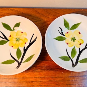 May include: Two white ceramic plates with a hand-painted floral design. Each plate features a yellow flower with red accents, green leaves, and black branches. The plates are set on a wooden surface.