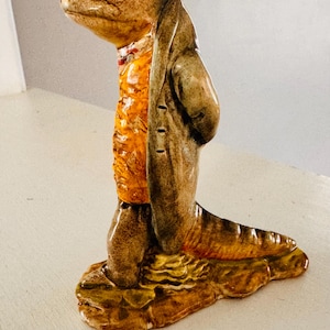 May include: A ceramic figurine of a lizard-like creature wearing a brown coat over an orange vest. The figurine has a detailed, textured appearance with a base that resembles a rock. The color palette includes shades of brown, orange, and yellow.