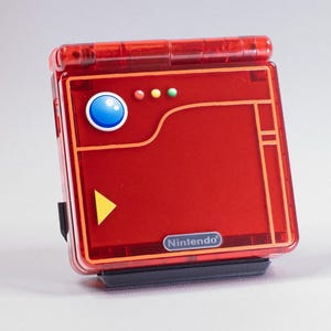 Op de afbeelding: Een rood draagbaar elektronisch apparaat met een transparante hoes. Het heeft een blauwe knop, een gele driehoek en drie kleine gekleurde lampjes. Het Nintendo-logo is zichtbaar. Het apparaat staat op een zwarte basis.