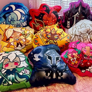 Puede incluir: Una colección de coloridas almohadas de felpa con forma de estrella con diseños de personajes de dibujos animados. Las almohadas muestran varios personajes en tonos de azul, rojo, amarillo, verde y rosa. Las almohadas están dispuestas sobre una colcha rosa.