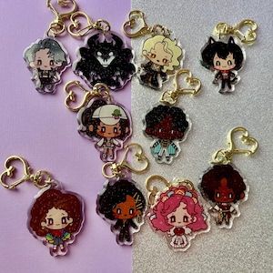 May include: A collection of acrylic keychains featuring colorful anime-style character designs. Each keychain has a gold-colored clasp. The characters have various hairstyles and outfits, with some holding accessories. The background is a mix of purple and silver.