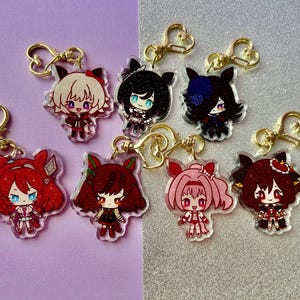Könnte beinhalten: Sammlung von sieben Anime-Acryl-Schlüsselanhängern mit goldfarbenen herzförmigen Verschlüssen. Jeder Schlüsselanhänger zeigt ein anderes Charakterdesign mit unterschiedlichen Haarfarben, Outfits und Gesichtsausdrücken. Der Hintergrund ist geteilt in Lila und Silber.
