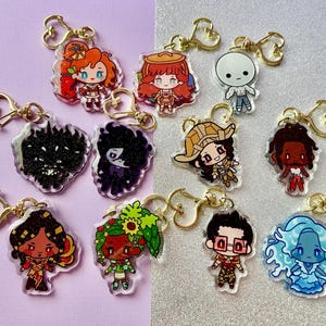 May include: A collection of acrylic keychains featuring various cartoon characters. Each keychain has a gold-colored heart-shaped clasp. The characters have different hair colors, outfits, and expressions, with some appearing to be fantasy-themed.