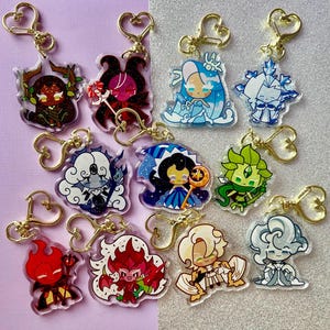 Op de afbeelding: Een verzameling karakter sleutelhangers met goudkleurige hartvormige sluitingen. Elke sleutelhanger heeft een ander cartoonpersonage in verschillende kleuren, waaronder rood, blauw, groen en wit. De sleutelhangers zijn gemaakt van helder acryl.