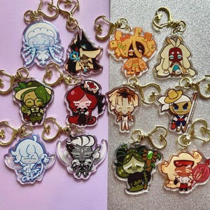 May include: A collection of twelve acrylic keychains featuring various cartoon character designs, each equipped with a gold-toned heart-shaped clasp. The characters display diverse color palettes including blue, green, red, orange, and black.