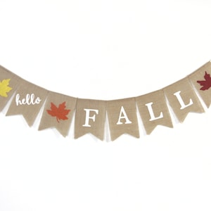 May include: A burlap banner with the words "hello fall" and a red, orange, and yellow leaf design. The banner is strung with white twine.