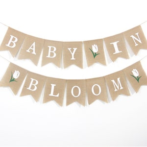 May include: A burlap banner with white letters spelling "BABY IN BLOOM" with a white tulip with a green stem on each end of the banner.