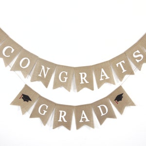 May include: A burlap banner with the words "CONGRATS GRAD" in white letters. The banner is decorated with black graduation caps with red tassels.