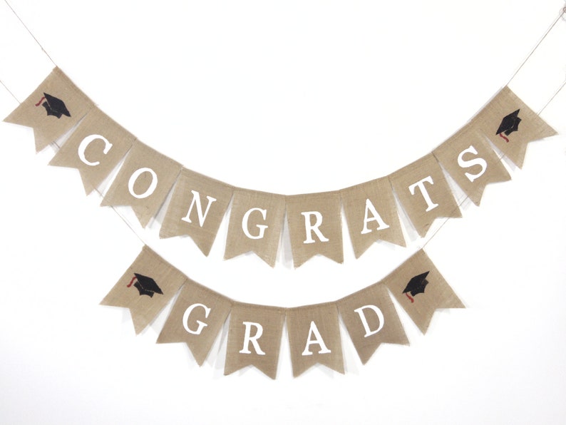 May include: A burlap banner with the words "CONGRATS GRAD" in white letters. The banner is decorated with black graduation caps with red tassels.