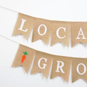 May include: A burlap banner with the words "LOCAL GROW" in white letters. The banner has a small orange carrot with green leaves on one of the flags.