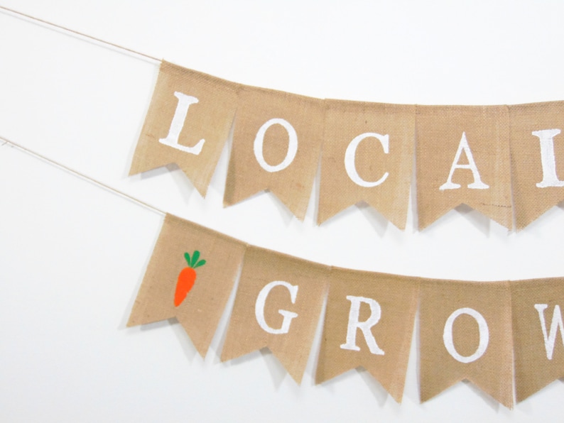 May include: A burlap banner with the words "LOCAL GROW" in white letters. The banner has a small orange carrot with green leaves on one of the flags.