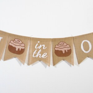 May include: A burlap banner with the words "Roll in the Dough" in white paint. The banner has two cinnamon roll illustrations.
