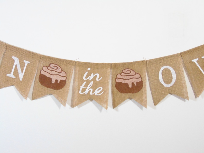 May include: A burlap banner with the words "Roll in the Dough" in white paint. The banner has two cinnamon roll illustrations.