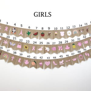 May include: A burlap banner with pink and white embroidered designs. The banner has 52 flags with numbers 1 through 52. The banner says "GIRLS" at the top.