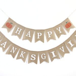 Op de afbeelding: Een banner van jute met de woorden "Happy Thanksgiving" in witte letters. De banner is versierd met twee oranje pompoenen met groene stelen.