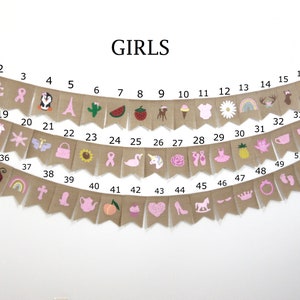 May include: A burlap banner with pink and white illustrations of various girls' themed items, including a unicorn, a flamingo, a crown, a teapot, a cupcake, a ballerina, a pineapple, a rainbow, a ladybug, a butterfly, a flower, a watermelon, a cactus, a Christmas tree, a rocking horse, a pair of ballet shoes, a shoe, a heart, a peach, a pumpkin, a bow, a teacup, a star, a llama, a purse, a sunflower, a ribbon, a cross, a boot, a deer, a bird, and an anchor. The banner is numbered 1 through 52.