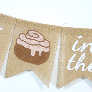 May include: A burlap banner with the words "Roll in the" and a brown and pink cinnamon roll graphic.