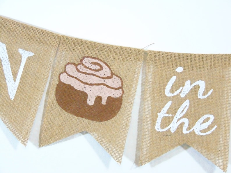 May include: A burlap banner with the words "Roll in the" and a brown and pink cinnamon roll graphic.