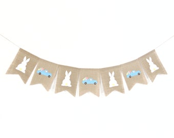 May include: A burlap banner with alternating white bunny and blue truck designs. The truck is carrying pastel colored eggs. The banner is strung with white twine.