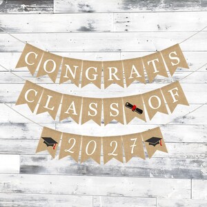 May include: A three-tiered burlap banner featuring white lettering that spells out CONGRATS CLASS OF 2027. The design includes a black and red diploma graphic replacing the letter O in OF, and two black graduation caps on the bottom row.