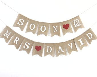 May include: A burlap banner with white text and red heart accents. The top banner reads "SOON TO BE" and the bottom banner reads "MRS DAVID". The banner is strung on a thin white string, perfect for a wedding or engagement celebration.