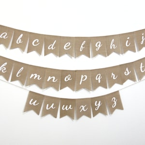 May include: A burlap banner with the alphabet in white cursive letters. The banner is strung on a white background.