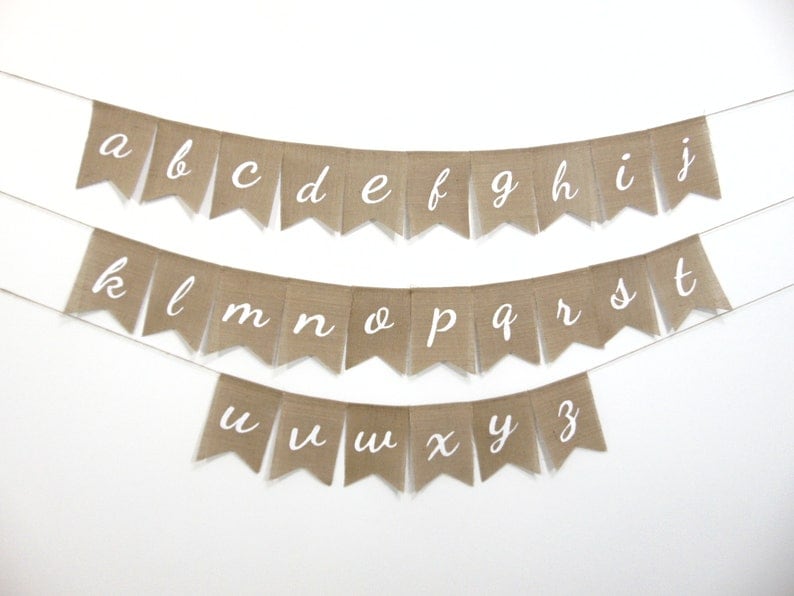 May include: A burlap banner with the alphabet in white cursive letters. The banner is strung on a white background.