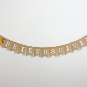May include: A tan burlap banner with white lettering spelling "FRIENDSGIVING". The banner features a decorative element with a yellow fruit and red and green accents. It is strung on a thin twine.