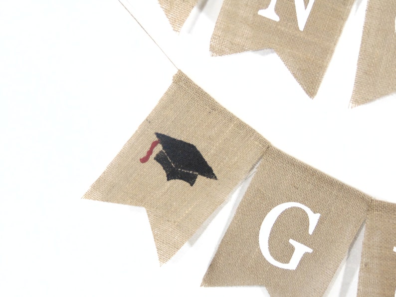 May include: A burlap banner with the word "GRAD" in white letters and a graduation cap graphic. The banner is perfect for a graduation party.