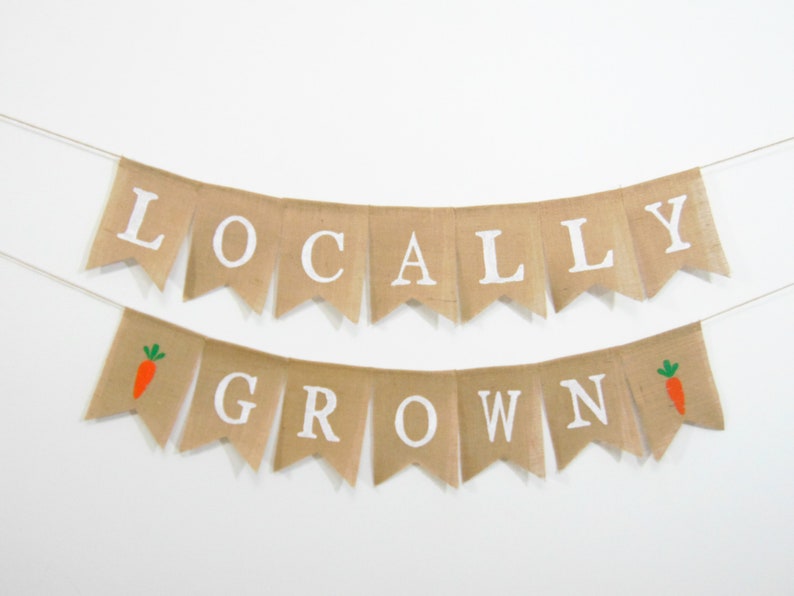 May include: A burlap banner with the words "LOCALLY GROWN" in white letters. The banner has two small orange carrots with green tops on either end.