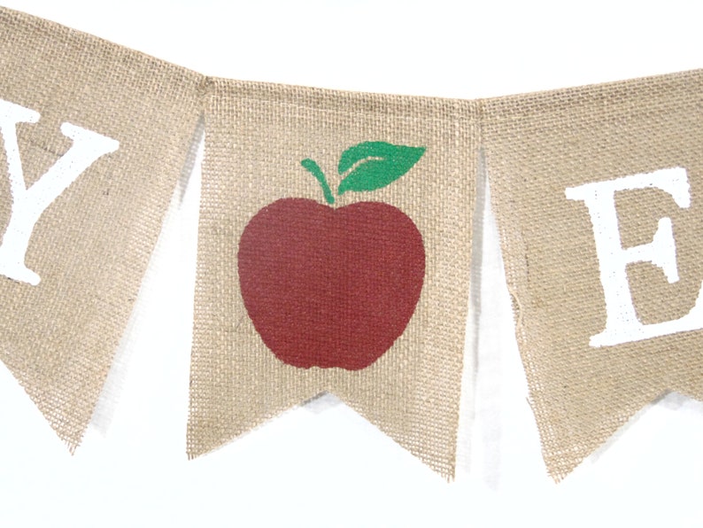 Puede incluir: Una pancarta de arpillera con las palabras "Happy" y "Fall" en letras blancas. La bandera del medio presenta una manzana roja con una hoja verde.