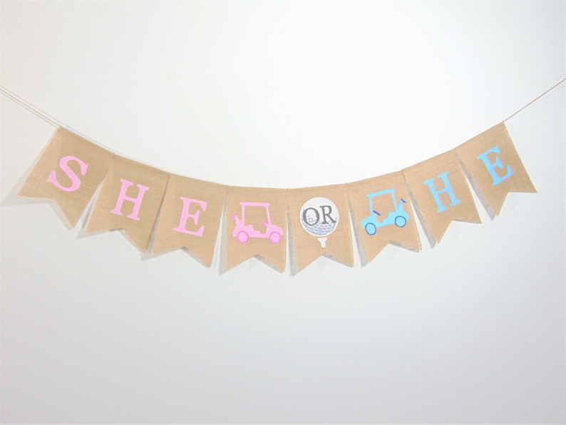 May include: A burlap banner with the words "SHE OR HE" in pink and blue letters. The banner also features a pink golf cart, a golf ball, and a blue golf cart.