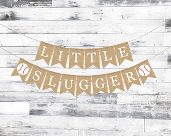 Peut inclure: Une bannière en toile de jute avec les mots "Little Slugger" en lettres blanches. La bannière est décorée de balles de baseball.